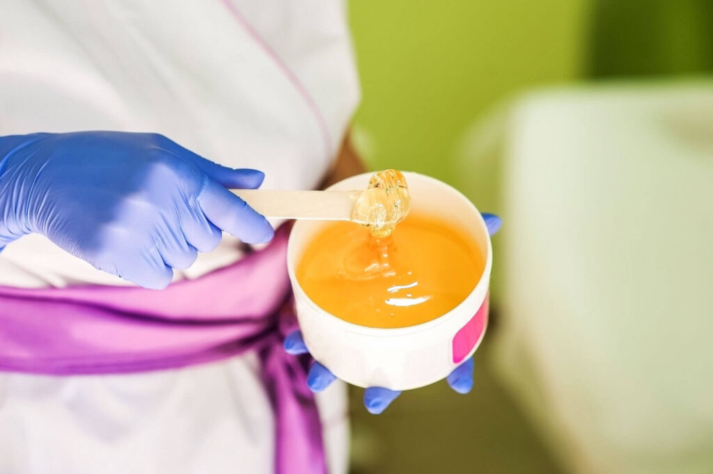 Blue gloves hold spatula with wax for depilation